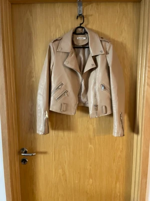 Beige skinnjacka från Cherry Koko - Snygg beige skinnjacka från Cherry Koko i bikerstil med silvriga dragkedjor och axelklaffar. Jackan har flera fickor med dragkedja och coola detaljer som ger en trendig look. Perfekt för dig som vill ha en stilren och modern jacka.