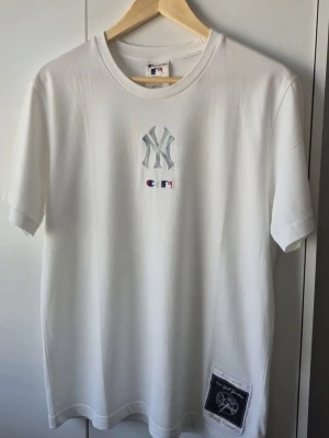 New York Yankees x Champion - Vit t-shirt från Champion med New York Yankees broderat på bröstet och officiell MLB-logga. Klassisk passform med rund hals och korta ärmar. Liten patch nertill på framsidan. Perfekt för dig som gillar sportig streetstyle.