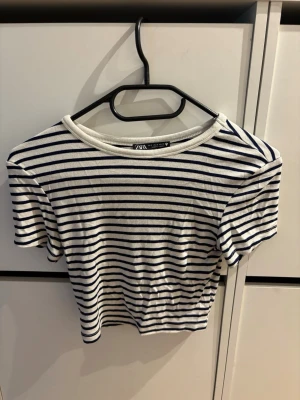 Randig topp från Zara i bomull - Snygg kortärmad topp från Zara i vitt med marinblå ränder. Klassisk rund halsringning och croppad passform. Tillverkad i mjuk bomull som känns skön mot huden. Perfekt för en avslappnad och trendig look.