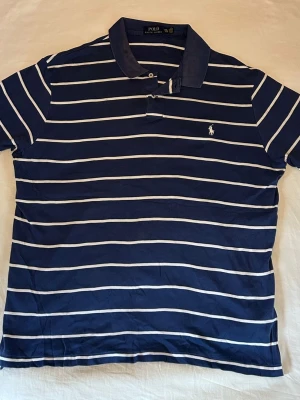Ralph Lauren | Piké | L (Står XXL men har L mått) - Mörkblå pikétröja från Ralph Lauren med stilrena ränder som ger en snygg och välklädd look. Den mörkblå färgen tillsammans med ränderna gör den både elegant och lite mer speciell. Broderad logga på bröstet och en bekväm passform som passar perfekt till både vardag och mer uppklädda tillfällen. Står XXL men passar som L, mått: Längd 71cm Bredd 60cm