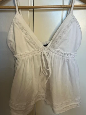 Vit linnetopp med spets från Gina Tricot - Supersöt vit linnetopp från Gina Tricot med smala axelband, spetsdetaljer och knyt framtill. Toppen har v-ringning och resår under bysten för en bekväm passform. Perfekt till sommaren och enkel att styla med jeans eller kjol.
