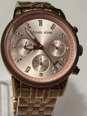 Guldig Michael Kors klocka - Stilren klocka från Michael Kors i guld med länkarmband och glittriga stenar på urtavlan. Klockan har flera små urtavlor, datumvisning och en snygg roséguldig ton på urtavlan. Perfekt accessoar för att lyfta din outfit.
