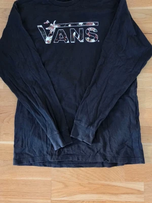 Svart långärmad tröja från Vans - Svart långärmad tröja från Vans i 100% bomull. Stor Vans-logga med blommigt mönster på bröstet. Klassisk passform med ribbade muddar vid ärmslut och hals. 