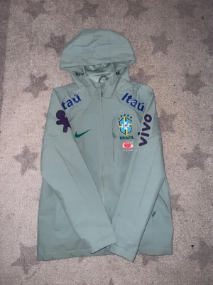 Nike Brazil turquoise windbreaker  - Snygg mintgrön vindjacka med huva från Hype the Detail. Jackan har coola mörklila tryck på båda ärmarna och stängs med dragkedja. Perfekt för blåsiga dagar och enkel att matcha med streetwear. Materialet är lätt och smidigt.