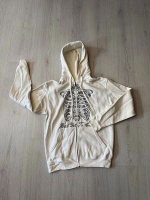 Rhinestone Hoodie - Säljer en vit hoodie med dragkedja och huva. På framsidan finns ett stort svart tryck av ett revben/skelett. Hoodien har fickor framtill och ribbade muddar. Perfekt för dig som gillar streetwear och unika prints. Har tyvärr en gul fläck på ärmen.
