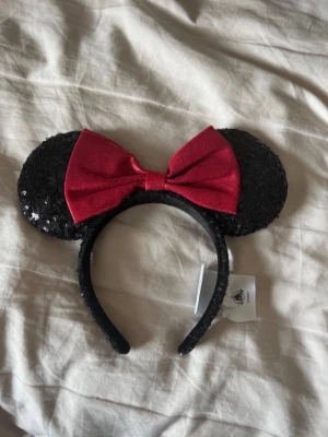 Disney Mimmi öron - Snyggt diadem från Disney med stora svarta paljettöron och en röd rosett i mitten. Perfekt accessoar för att sticka ut och ge en lekfull touch till din outfit. Diademet är täckt av glittriga paljetter och har en klassisk Minnie Mouse-stil.