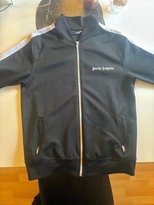 Svart zip-hoodie från Palm Angels - Svart hoodie från Palm Angels med vit dragkedja och vita ränder längs ärmarna. Klassisk logga på bröstet och två fickor med dragkedja. Hög krage och sportig look, perfekt för streetwear-stilen.
