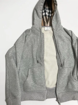 Grå Zip-hoodie med rutigt foder - Storlek S - Säljer denna tunga zip-hoodie i gråmelerat utförande. Materialet är en kraftig bomullsfleece som känns rejäl och håller formen bra. Huvan har det klassiska rutiga fodret på insidan och bröstet pryds av en broderad logotyp i matchande ton. Tröjan har detaljer i högpolerat silver och graverade metallavslut på dragsnörena. Alla invändiga etiketter sitter kvar. Den är i mycket gott skick utan noppor eller slitage. Skicka ett meddelande vid frågor eller intresse. 