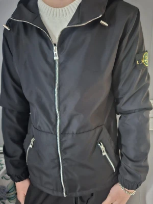Svart vindjacka från Stone Island - Svart vindjacka från Stone Island med huva, hel dragkedja framtill och två snedställda fickor med dragkedja. Jackan har elastiska muddar vid ärmslut och klassisk logotyp-patch på vänster ärm. Perfekt för en stilren och sportig look.