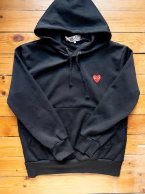 Svart hoodie från Comme des Garçons Play - Svart hoodie med huva och dragsko från Comme des Garçons Play. Klassisk design med röd hjärtlogga med ögon broderad på bröstet. Mjuk bomullskvalitet, ribbade muddar och känguruficka framtill. Perfekt för en stilren och avslappnad look.