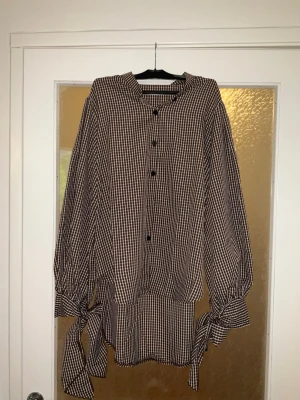 Rutig blus med knytband i ärmarna - Snygg blus i svartvitt rutigt mönster med långa ärmar och knytband vid ärmsluten. Blusen har knappar framtill och en lös, avslappnad passform. Perfekt för dig som gillar en trendig och bekväm stil.