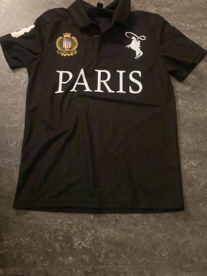 Svart Paris t-shirt med tryck - Svart t-shirt med vit text 'PARIS' framtill, broderad emblem i guld och lila samt en vit hästlogga på bröstet. Klassisk krage och korta ärmar. Perfekt för dig som gillar streetstyle och vill sticka ut med snygga detaljer.