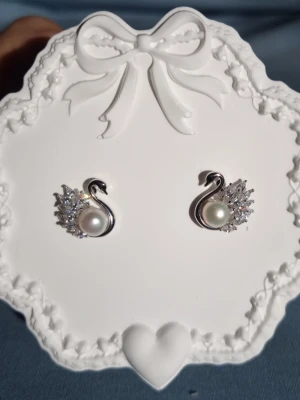 Eternal Love (silver 925 stift)  - Svanar är en symbol för kärlek, trohet och evighet – de representerar starka band och livslång samhörighet. Smycket är dekorerat med en sötvattenspärla, CZ-diamanter som reflekterar ljuset och ger en exklusiv lyster. Tillverkad i Korea.                                   Material: Rhodium, Stift i 925 silver (stämplat), sötvattenspärlor, CZ-diamant Storlek: längd ca 1,5 cm Bredd ca 1,5 cm -Inga prisförslag, tack                                        - Varan packas noggrant och säkert 