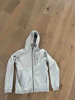 Ljusgrå hoodie från Peak Performance - Snygg ljusgrå hoodie med dragkedja från Peak Performance. Tröjan har huva med dragsko, två fickor framtill och en diskret logga på bröstet. Perfekt för en avslappnad och sportig stil.