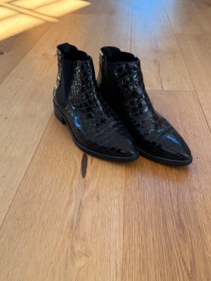 Vagabond Boots - Snygga svarta Chelsea boots med glansig croco-imitation och spetsig tå. Resår på sidorna för enkel påtagning och låg klack. Perfekta för att lyfta din outfit med en trendig touch.