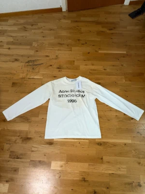Vit långärmad t-shirt Acne Studios - Vit långärmad t-shirt från Acne Studios med svart text 'Acne Studios STOCKHOLM 1996' på bröstet. Klassisk rak passform och rund halsringning. Tillverkad i mjuk bomull, perfekt för en clean och stilren look.