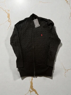 Svart kabelstickad ziptröja Polo Ralph Lauren - Kabelstickad svart ziptröja från Polo Ralph Lauren med röd broderad logga på bröstet. Tröjan har ribbade muddar, hög krage och dragkedja framtill. Perfekt för en stilren och klassisk look.