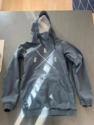 Blå vindjacka från Dope med huva - Snygg blå vindjacka från Dope med stor vit logga på bröstet. Jackan har huva med dragsko, magficka och elastiska muddar vid ärmslut och nederkant. Materialet är vattenavvisande och perfekt för blåsiga dagar. Aldrig använd.