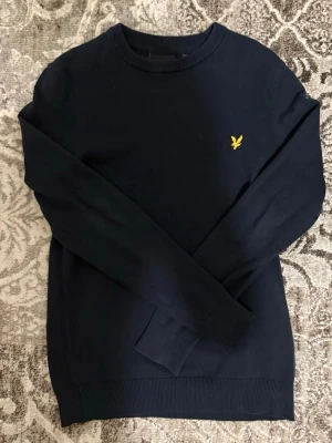 Mörkblå tröja från Lyle & Scott - Klassisk mörkblå tröja från Lyle & Scott med rund halsringning och den ikoniska gula örnen broderad på bröstet. Tröjan har långa ärmar och ribbade muddar vid ärmslut och nederkant. Perfekt för en stilren och enkel look.