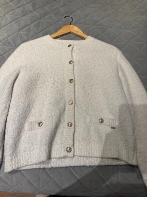 Vit kofta från Holly & Whyte - Säljer en fluffig vit kofta från Holly & Whyte by Lindex. Koftan har rund hals, dekorativa guldknappar framtill och på fickorna samt två fickor fram. Perfekt för dig som gillar en stilren och mysig look.