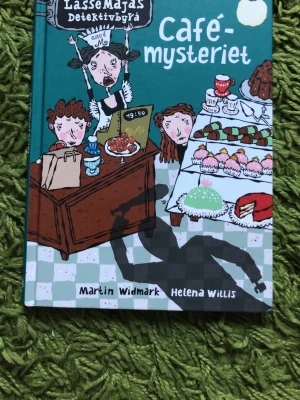 LasseMajas Detektivbyrå: Cafémysteriet - En färgglad och illustrerad bok i serien LasseMajas Detektivbyrå, skriven av Martin Widmark och illustrerad av Helena Willis. Boken har hårda pärmar i turkos med tecknade figurer och bakverk på omslaget. Perfekt för dig som gillar mysterier och spännande berättelser.