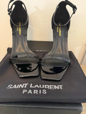 Svarta lackpumps från Saint Laurent - Snygga svarta pumps i lack från Saint Laurent med öppen tå, smal rem över tårna och justerbar ankelrem. Klacken är i metall med YSL-logga, vilket ger en lyxig och trendig look. Perfekta för dig som vill sticka ut med exklusiva detaljer.
