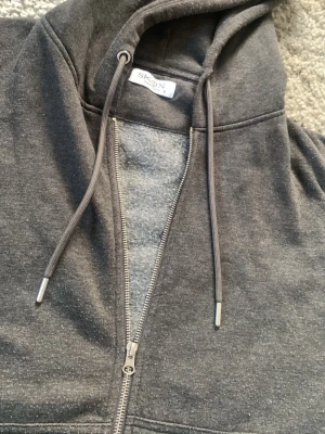 Mörkgrå zip-hoodie från SKØN - Mysig mörkgrå hoodie från SKØN Copenhagen med dragkedja framtill och justerbar huva med snören. Insidan är mjukt borstad för extra komfort. Perfekt för chill dagar eller när du vill ha en avslappnad look nu till våren.