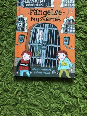 Fängelsemysteriet - LasseMajas Detektivbyrå - En färgglad och illustrerad bok ur serien LasseMajas Detektivbyrå, skriven av Martin Widmark och illustrerad av Helena Willis. Omslaget är orange med tecknade figurer och ett fängelse. Perfekt för unga som gillar mysterier och spännande äventyr.