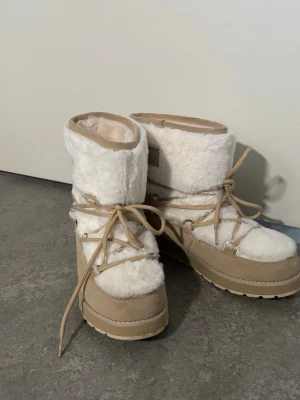 Vita moon boots från Luhta - Säljer ett par snygga vita moon boots från Luhta med beige detaljer och snörning runt om. Bootsens ovandel är i mjuk fuskpäls och nederdelen i mockaimitation. Perfekta för kalla vinterdagar och ger en riktigt cozy vibe. Endast använd en gång