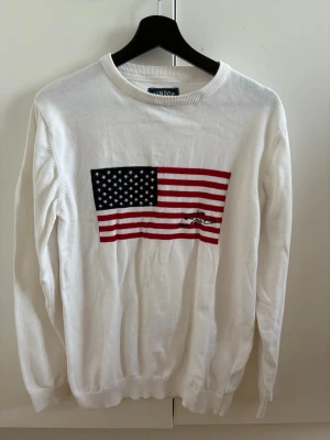 Vit stickad tröja med USA-flagga - Vit stickad tröja med en stor amerikansk flagga broderad på bröstet. Tröjan har rund halsringning och långa ärmar. 