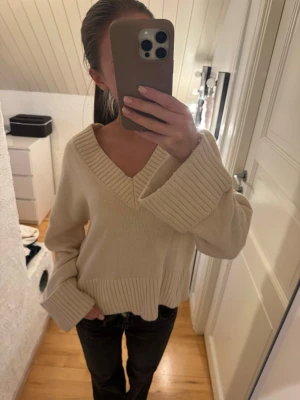 Beige stickad tröja med v-ringning - Mysig beige stickad tröja med bred v-ringning och ribbade detaljer vid hals, ärmslut och nederkant. Tröjan har en avslappnad passform och vida ärmar.