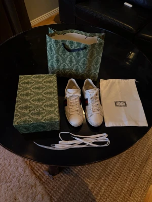 Gucci vita sneakers med röd/blå rand - Säljer ett par klassiska vita sneakers från Gucci med den ikoniska röd-blåa sidoremsan. Skorna är i läder och har vita snören. Medföljer originalkartong, dustbag och extra skosnören. Perfekta för dig som vill ha en stilren och lyxig look.