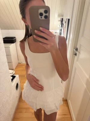 Vit kort klänning med volang - Supersöt vit kort klänning med fyrkantig ringning och flera lager volanger nedtill. Klänningen är ärmlös och har en figurnära passform.