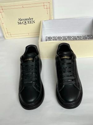 Svarta Alexander McQueen sneakers - Stilrena helsvarta sneakers från Alexander McQueen i slät läder med tjock sula och svarta skosnören. Diskret guldtext på plösen och hälen. Klassisk low-top modell med minimalistisk design och perforerade detaljer på sidan.