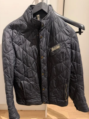 Svart quiltad jacka från matchless - Snygg svart quiltad jacka från matchless med  hög krage, tryckknappar och dragkedja. Jackan har ett klassiskt matchless-märke på bröstet och praktiska fickor med dragkedja. Perfekt för dig som vill ha en stilren och tidlös look.