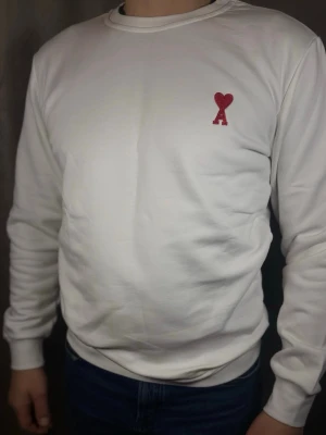 Vit sweatshirt från Ami Paris - Vit sweatshirt från Ami Paris med röd broderad logga i form av ett hjärta och bokstaven A på bröstet. Klassisk rund halsringning och långa ärmar. Tillverkad i mjuk bomull för en bekväm känsla.