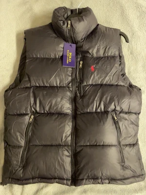 Svart dunväst från Polo Ralph Lauren - Snygg svart dunväst från Polo Ralph Lauren med hög krage och dragkedja framtill. Västen har två snedställda fickor med dragkedja och den klassiska röda loggan broderad på bröstet. Perfekt för lager-på-lager och riktigt varm tack vare dunfyllningen.