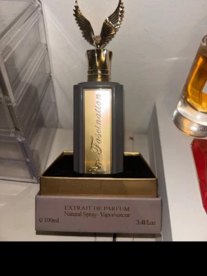 Fascination Extrait de Parfum 100ml - Lyxig parfymflaska i svart och guld med elegant rektangulär form och en unik kork med guldfärgade vingar. Flaskan rymmer 100 ml och har en exklusiv design som verkligen sticker ut på parfymhyllan. Perfekt för dig som gillar statement-detaljer.