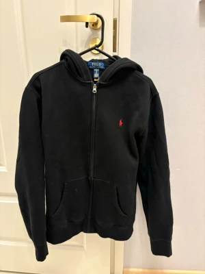 Svart hoodie från Polo Ralph Lauren - Klassisk svart hoodie med dragkedja från Polo Ralph Lauren. Har huva, kängurufickor och den ikoniska röda loggan broderad på bröstet. Tillverkad i mjukt bomullsmaterial som är skönt att bära. Perfekt för en avslappnad och stilren look.