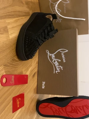 Svarta Christian Louboutin sneakers - Snygga svarta sneakers från Christian Louboutin med mattsvarta nitar på tån och klassisk röd sula. Skorna har en låg silhuett, svarta skosnören och är tillverkade i mocka och läder. Perfekta för dig som vill sticka ut med exklusiv design.
