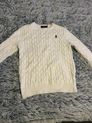 Vit kabelstickad tröja Polo Ralph Lauren - Kabelstickad vit tröja från Polo Ralph Lauren med rund halsringning och klassisk marinblå logga broderad på bröstet. Tröjan har ribbade muddar vid ärmslut och nederkant och är gjord i ett mjukt material som känns lyxigt.