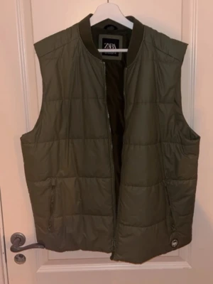 Olivgrön dunväst från Zara - Snygg olivgrön dunväst från Zara med quiltad design och dragkedja framtill. Västen har två fickor med dragkedja och ribbad krage. Perfekt för lager på lager och ger en clean look till din outfit.