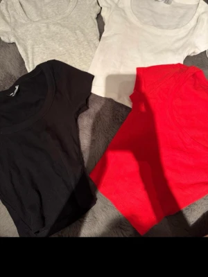 Fyra bas-t-shirts i olika färger - Fyra enkla t-shirts i svart, grått, vitt och rött. Alla har rund halsringning och korta ärmar, perfekta att matcha till olika outfits. Materialet känns mjukt och stretchigt, troligen bomull eller bomullsblandning.