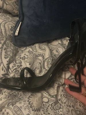 Svarta YSL pumps med logoklack - Säljer ett par svarta pumps från Saint Laurent med öppen tå och tunn rem över foten. Klacken är formad som YSL-loggan vilket ger en unik och lyxig look. Skorna är i blankt läder och har en elegant siluett som verkligen sticker ut. Skorna är helt oanvända,  lägger ut dom för ett billigt pris då kvitto inte finns kvar❤️