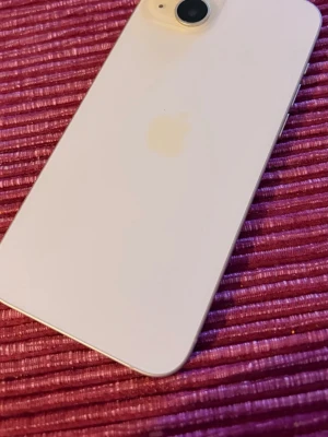 Apple iPhone mobiltelefon 128 GB - Säljer en vit iPhone med stilren design och rundade hörn. Baksidan är i glas och har en enkel kamera med blixt i hörnet. Apple-loggan syns diskret på baksidan. Framsidan har en stor skärm med tunna kanter, perfekt för sociala medier och appar.