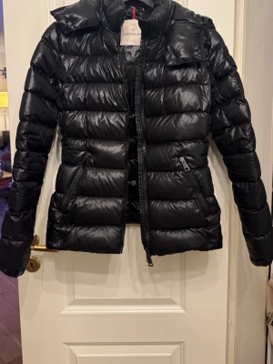 Svart dunjacka från Moncler - Snygg svart dunjacka från Moncler med glansig finish och quiltad design. Jackan har dragkedja framtill, två snedställda fickor med dragkedja och en Moncler-logga på ärmen. Den är köpt 2020 och är köpt på NK. Kvitto medföljer samt lådan om man vill ha den med