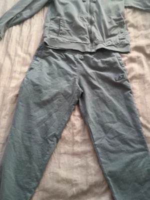 Grått EA7 loungewear-set - Säljer ett grått loungewear-set från EA7 med matchande hoodie och byxor. Hoodien har dragkedja, fickor och huva med snören. Byxorna har resår i midjan och EA7-logga på benet. Perfekt för chill dagar hemma.