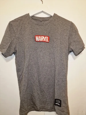 Grå Marvel t-shirt i bomull - Snygg grå t-shirt från Marvel med klassisk röd och vit Marvel-logga på bröstet. Perfekt för dig som gillar superhjältar och vill ha en enkel men cool look.
