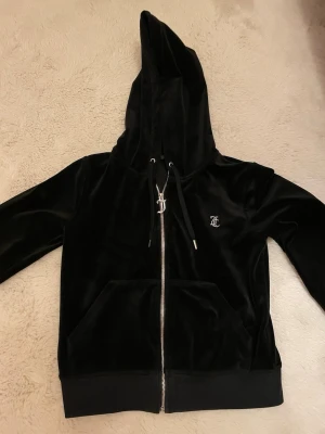 Svart velour hoodie från Juicy Couture - Snygg svart hoodie i mjuk velour från Juicy Couture med dragkedja och glittrande J-dragkedja. Klassisk huva, känguruficka och liten Juicy Couture-logga broderad på bröstet. Perfekt för en trendig och bekväm look. Förekommer defekter som framkommer på bilden. 