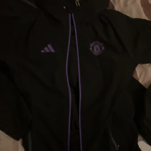 Manchester United träningsset Adidas - Säljer ett svart träningsset från Adidas x Manchester United med lila detaljer. Setet består av en hoodie med dragkedja och byxor, båda med klubbens logga och Adidas-märke i lila. Perfekt för fotbollsträning eller chill.